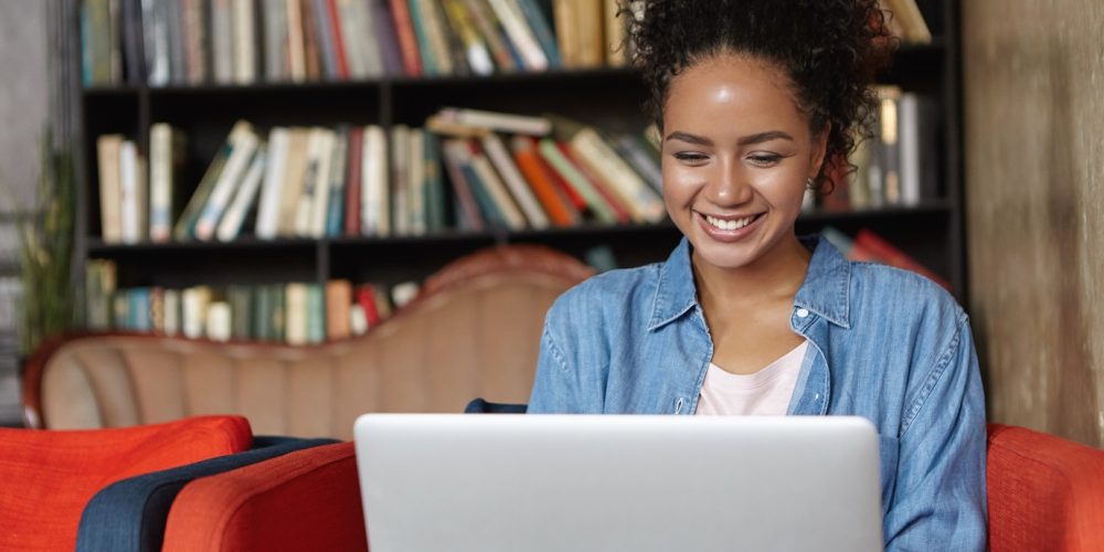 People, technology and education concept. Happy pretty dark-skinned female in jean shirt sitting at sofa using laptop, typing something and browsing internet while being in reading hall or library.